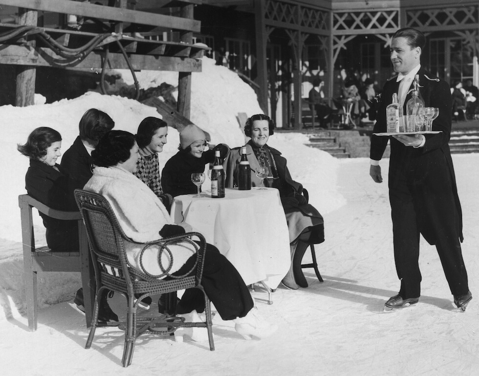 A waiter brings, ice skating, a tray of gin and tonic to Lady Scarsdale (right) on holiday with her daughters in St Moritz, Switzerland